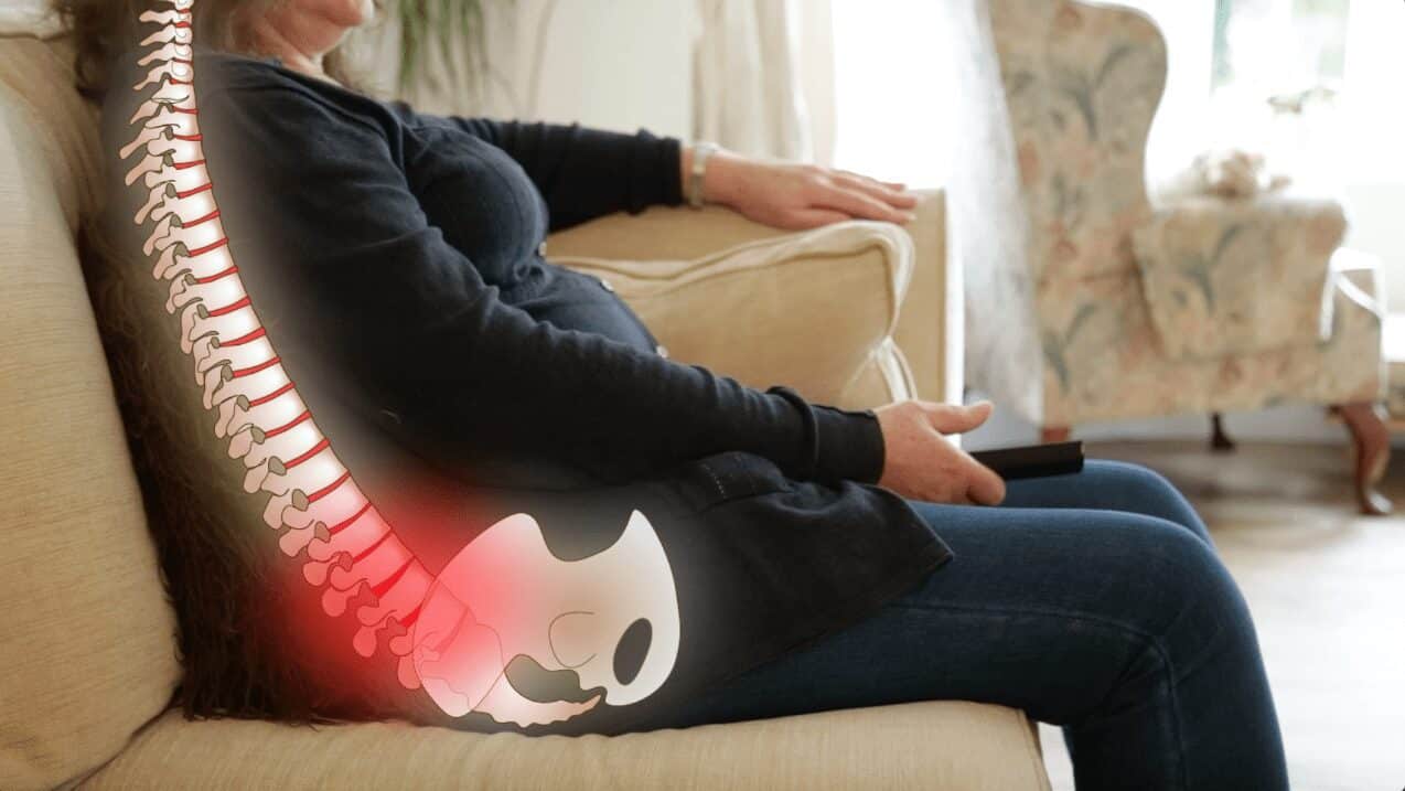 A lady is sat side on she is sat on a sofa slouching. Overlaid is an image of what is going on in her spine with a red shading area to show pressure into her lower back