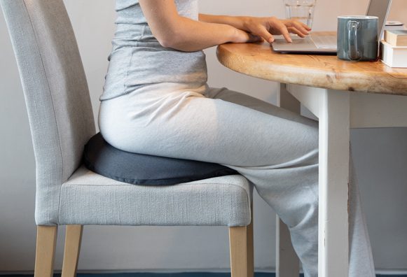 A side on image of a lady wearing grey casual sports wear she is sat facing to the right although you can only see part of her torso and her legs. She is sat at the laptop with water in the background and a hot drink in the foreground. The table has a wooden top to it. She is sat on a dining room chair which is also grey and she has the Putnams coccyx ring on there to help relieve her coccyx pain whilst she is sat working