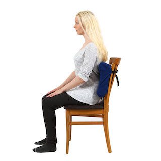 A lady in profile sat on a wooden chair she is facing to the left. She has blonde hair a grey t-shirt and black trousers. She is sat against the Royal Rest Comfort plus cushion which is against the back of the chair