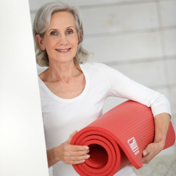 On the left of the image is an older lady with grey/white hair to just abover her shoulders. She is wearing white and smiling at the camera tucked under her left arm she is holding a red rolled up Sissel gym mat which she is supporting at the front with her right hand.