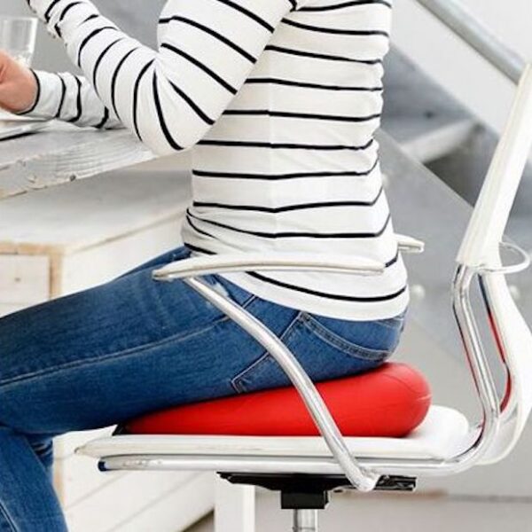 The whole scene is very white which includes the stairs in the background, walls, table a lady is working at and chair. She is sat side on working at a laptop, glass of water in the background. She is wearing blue jeans and a white top with black stripes, she is sat on a red Sitfit plus which really stands out.