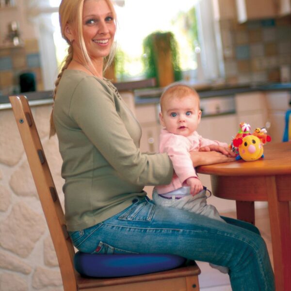 A lady is sat side on to the camera smiling she has a young baby on her lap who is also looking at the camera. There is a wooden circular table in front of them and there is a toy on the table you can just see that it is fluffy and yellow with black eyes and a pink nose. The lady is sat on a blue sitfit plus. The image behind is blurred you can make out that it is a kitchen