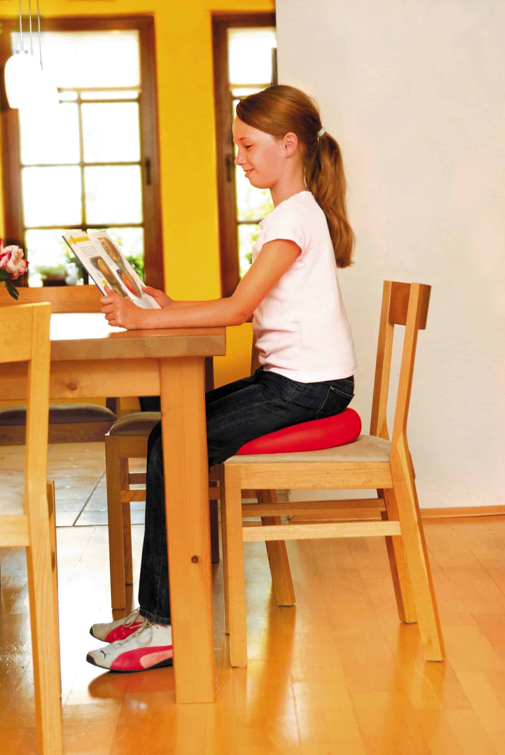 A child is sat at a wooden dining room table reating a book. She is sat on a red sitfit plus inflatable cushion