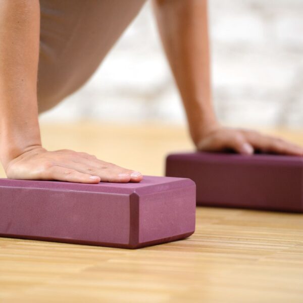 A pair of burgundy yoga blocks side by side. You can just make out that someone is possibly doing a cobra Yoga pose or maybe an upward dog pose. The palms of their hands are placed on top of each block.