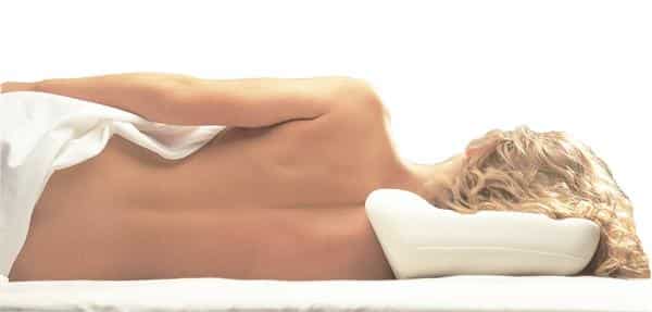 A lady with long blonde hair is facing away from the camera lying on her right hand side. She is sleeping on a Putnam's memory foam pillow. The bed sheet is moved away so that you can see her spine which is perfectly aligned due to the support of the mattress topper and the Putnam's pillow itself.