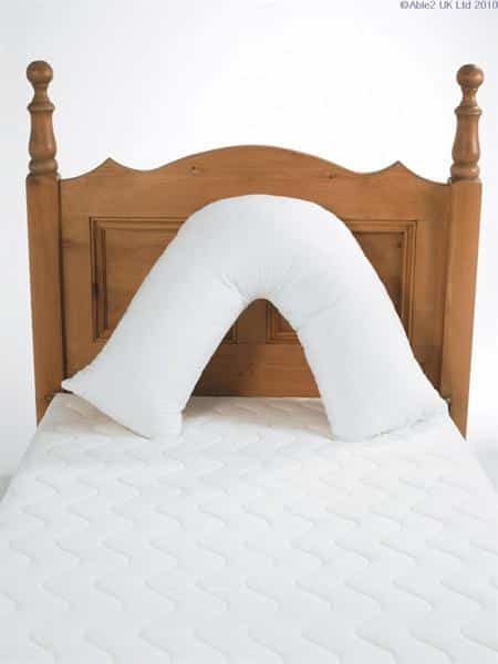 A Harley V shaped pillow is placed in front of a pine headboard. The Harley V shaped pillow is white and placed like an inverted v so there are two parts that are in contact with the surface you are sitting on and then it arches around so the top of it is supporting your upper and middle back.
