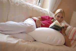 A lady who is heavily pregnant is holding her belly. She is lying on her left side enjoying reading a book whilst benefitting from the support of a Sissel comfort cushion which is also between her knees and supporting her pregnant belly.