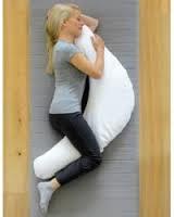 A blonde haired lady is lying on her side with a pregnancy support cushion. The pregnancy support cushion is white and in a half moon shape which is wider in the middle. She is resting her head on the upper section and has the lower part placed between her legs.