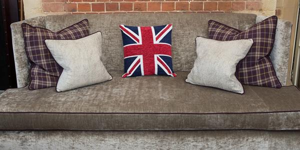 On a grey/silver velvet sofa are an array of scatter cushion in the centre a Union Jack cover. Either side is a purple tartan cushion at the back with smaller silvery sequin covered cushion.