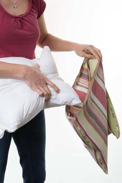 The model is holding a stripy cushion cover in their left hand so it is hanging down. In her right hand she has a Sittingwell back support cover. Her hand is spread so that she has pulled the two sides of the cushion together to make it smaller so that you can then easily slide it inside of the cushion cover to them zip it up and disguise it as any other cushion in your home.