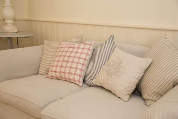 This is an image of four different cushions laid out side by side on the sofa they're light coloured cream/beige. The second cushion on the left has red horizontal and the one on the far right has vertical lines and the one in front of that has a pattern. One of these cushions has a Sittingwell back support cushion in it. The main aim of the photo is to highlight there is no need to have a obvious looking orthopaedic and medical looking back support in your home. From the image you can't tell where the back support is on the sofa.