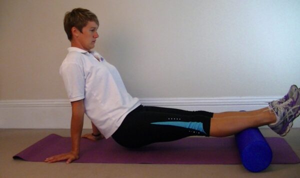 Lorna is wearing a white t-shirt and black jogging trousers and trainers she is on a purple gym mat with her hands behind her back. She has a blue foam roller placed behind her calf muscles so she is lifting herself slightly off the ground with her hands so that she can get the benefit of the calf massage.