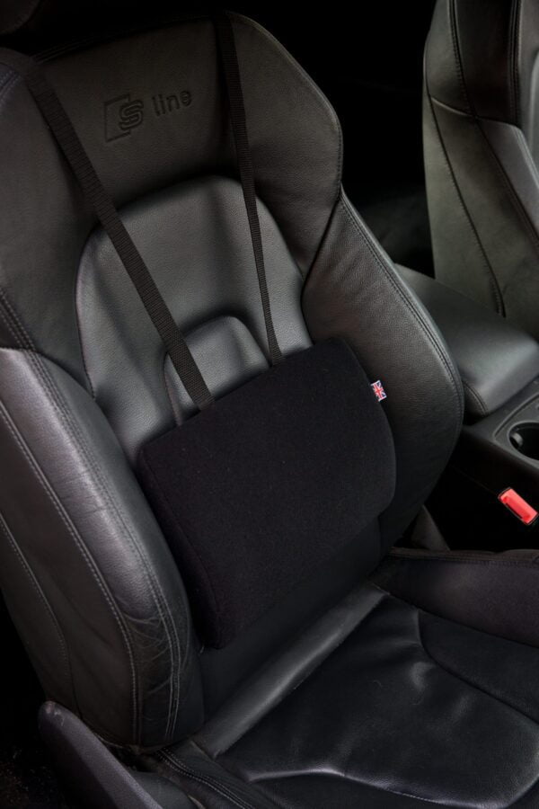 There is a black leather car seat which is facing towards the bottom right of the photo. The photo is zoomed in so you can see the details of the back support. On it is a black velour covered Duo car seat support. You can see that it is big enough for your lower back area and it hangs from a black strap that is placed over the top of the car seat and held in place by the headrest. The support itself sits flush against the back of the car seat.