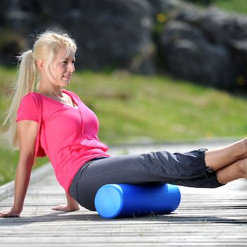 A lady with her legs on a foam roller sowing how you can use it for self-massage. She is outdoors although the background is blurred you can see it is a lovely sunny day and you can see there is a grassy slope in the background and maybe a stone wall or a mound of stones