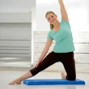 a lady is a light green top and possibly navy leggings doing a kneeling side stretch to the right with her left arm raised above her head. Her left knee is on the balance pad and her right leg stretched out to the side straight with her foot on the floor.