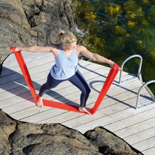 A blonde woman wearing blue leggins and a pale blue strappy top is standing in a lunge postition on a wooden platfrom wich is on rocks with metal steps on the right down to the sea. She has her felt leg forward and left arm stretched out in front and right arm stretched out behind her. She has a red resitance band wrapped round her left hand and it goes down under her left and right foot back up to where she is also holding it in her right hand putting tension throughout the band