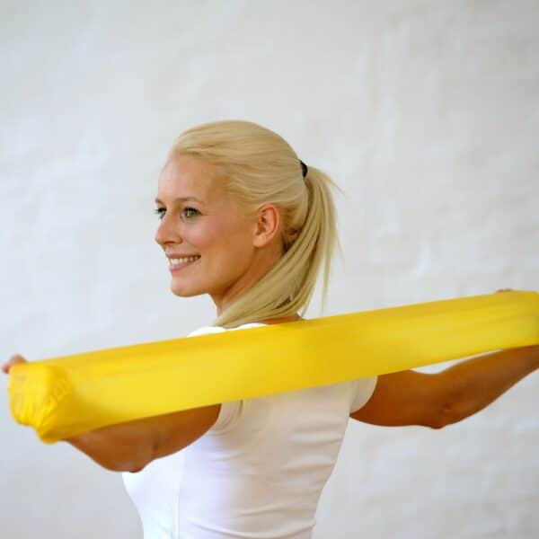 Standing in front of a white wall, a blonde lady in a white t-shirt has her back to the camera with her head truned tot he left. Her arms are out stretched horizontal to the floor and she is holding a yellow fun & active band on a stretch between her left and right hands.