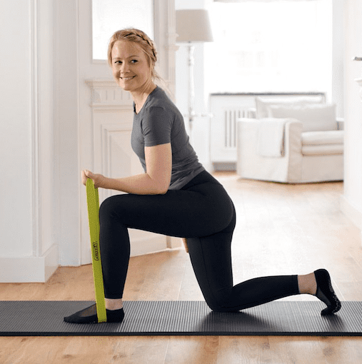 A lady kneeling a smiling towards the camera with a resistance band looped around her feet and holding it in her hand depicting that she will be doing a bicep strengthing exercise.