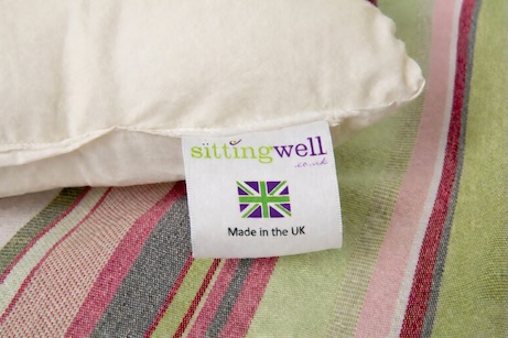 You can see the corner of the Sittingwell back support which is placed on a striped cushion cover. The image clearly shows the product label which has Sittingwell.co.uk written at the top, below is a Union Jack and below that the words made in the UK.