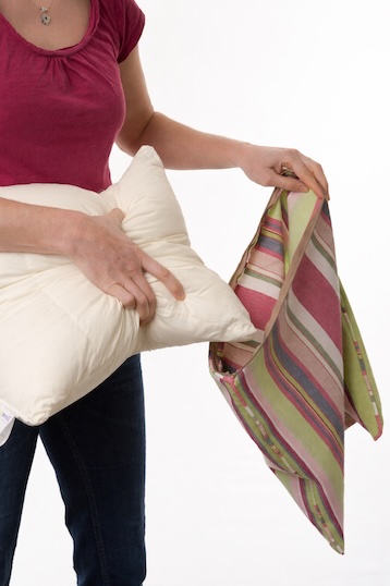 A lady has a Sittingwell Cushion In her right hand and gripping it to put into a stripy cushion cover. Her hand is spread so that she has pulled the two sides of the cushion together to make it smaller so that you can then easily slide it inside of the cushion cover to them zip it up and disguise it as any other cushion in your home.